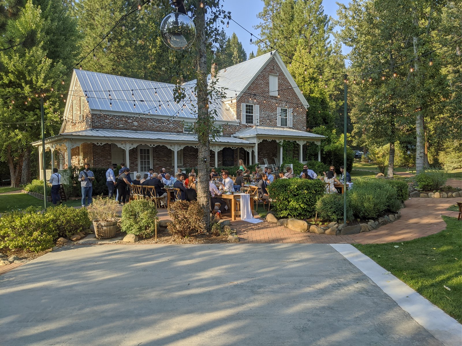Guests at dinner tables under the trees in front of the brick house at Twenty Mile House