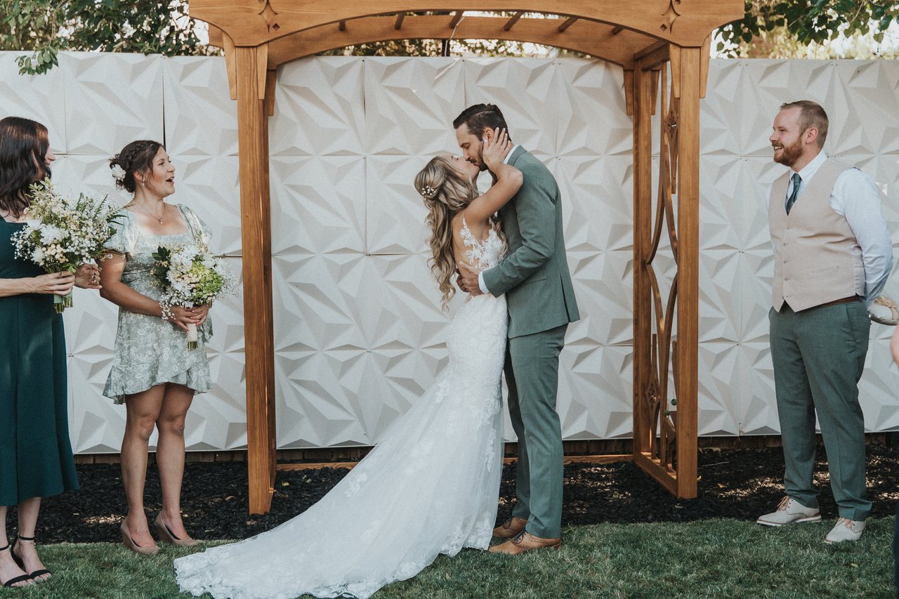 Lexy and Jeremy kiss under the arbor as the bridal party looks on