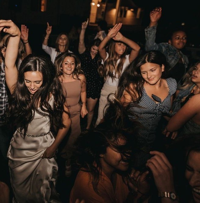Lindsey dances with her hand up in her satin dress, surrounded by friends on the dance floor at night