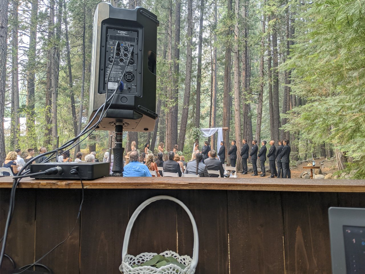 A forest wedding ceremony under tall pines at Twenty Mile House, seen from the DJ booth