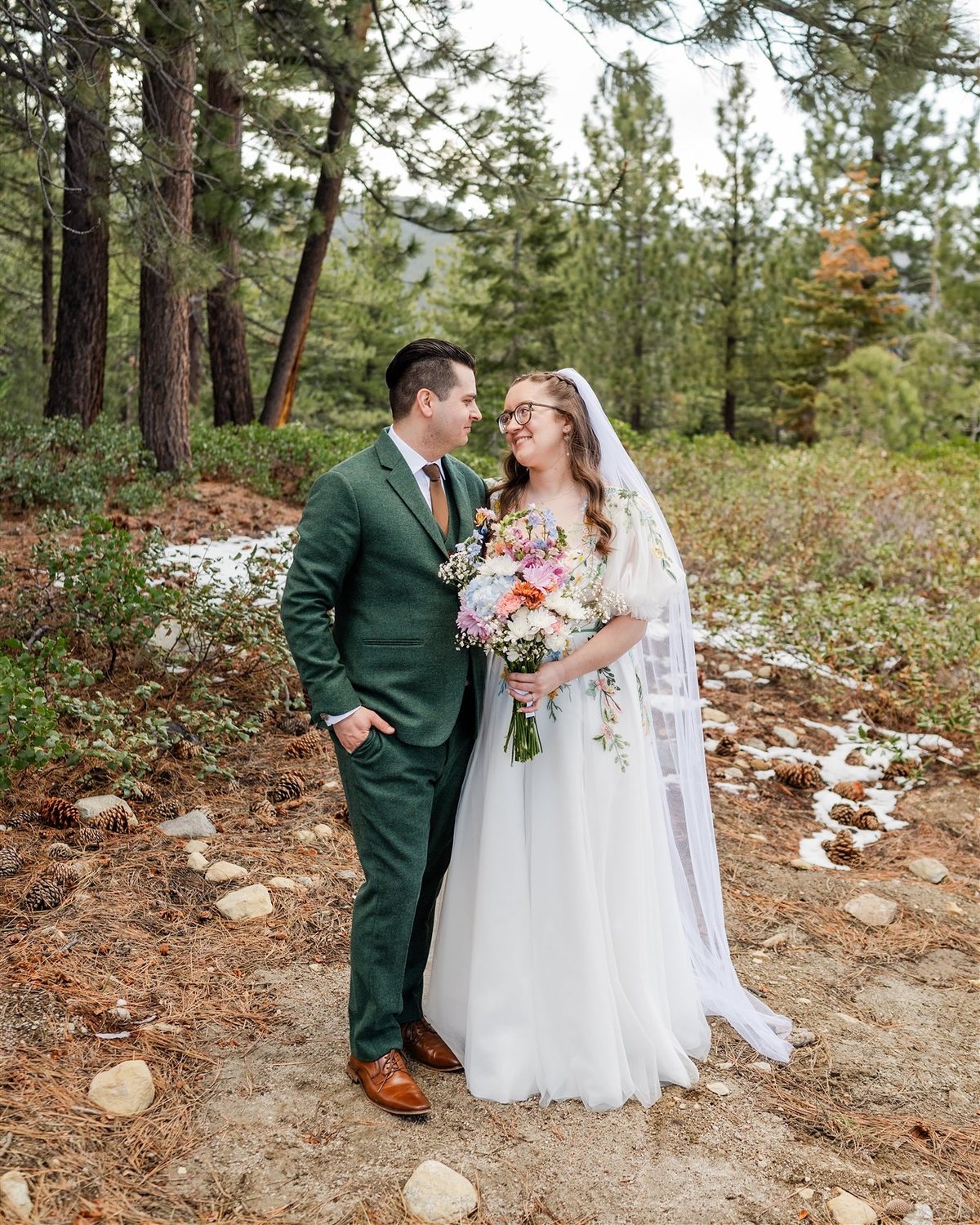 Madison and Adam stand together in the pines with patches of snow behind them