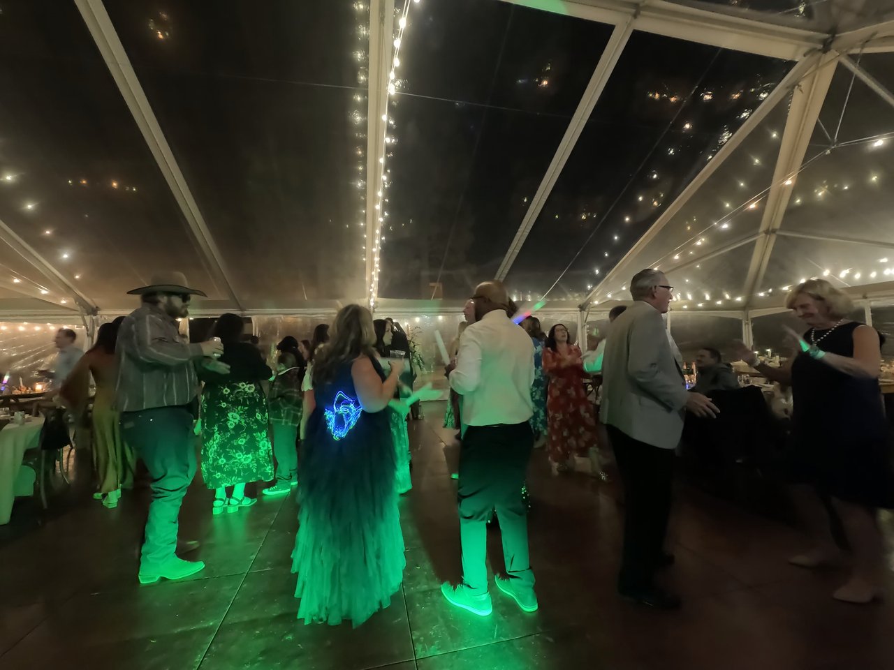 Guests dance with glow sticks under green lights and string lights in the clear tent