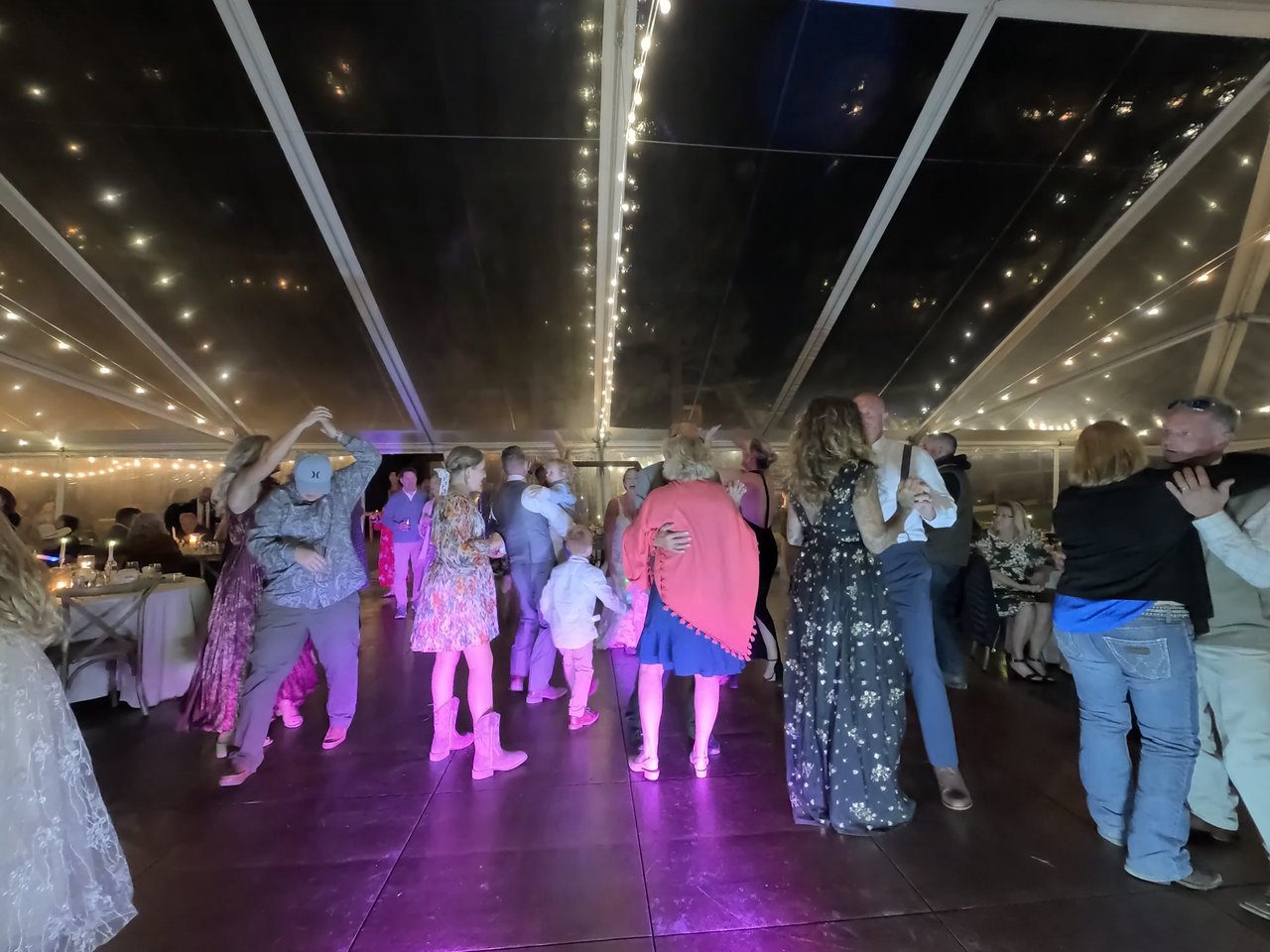A crowded dance floor under pink lights and string lights in the clear tent