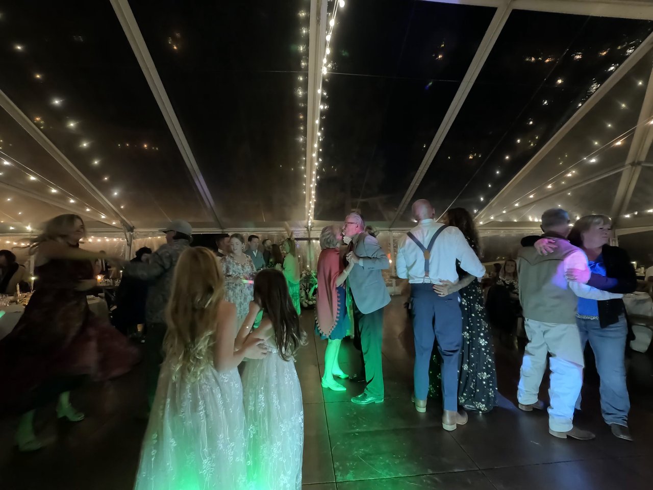 Two flower girls in lace dresses dance together as couples spin around them under the string lights