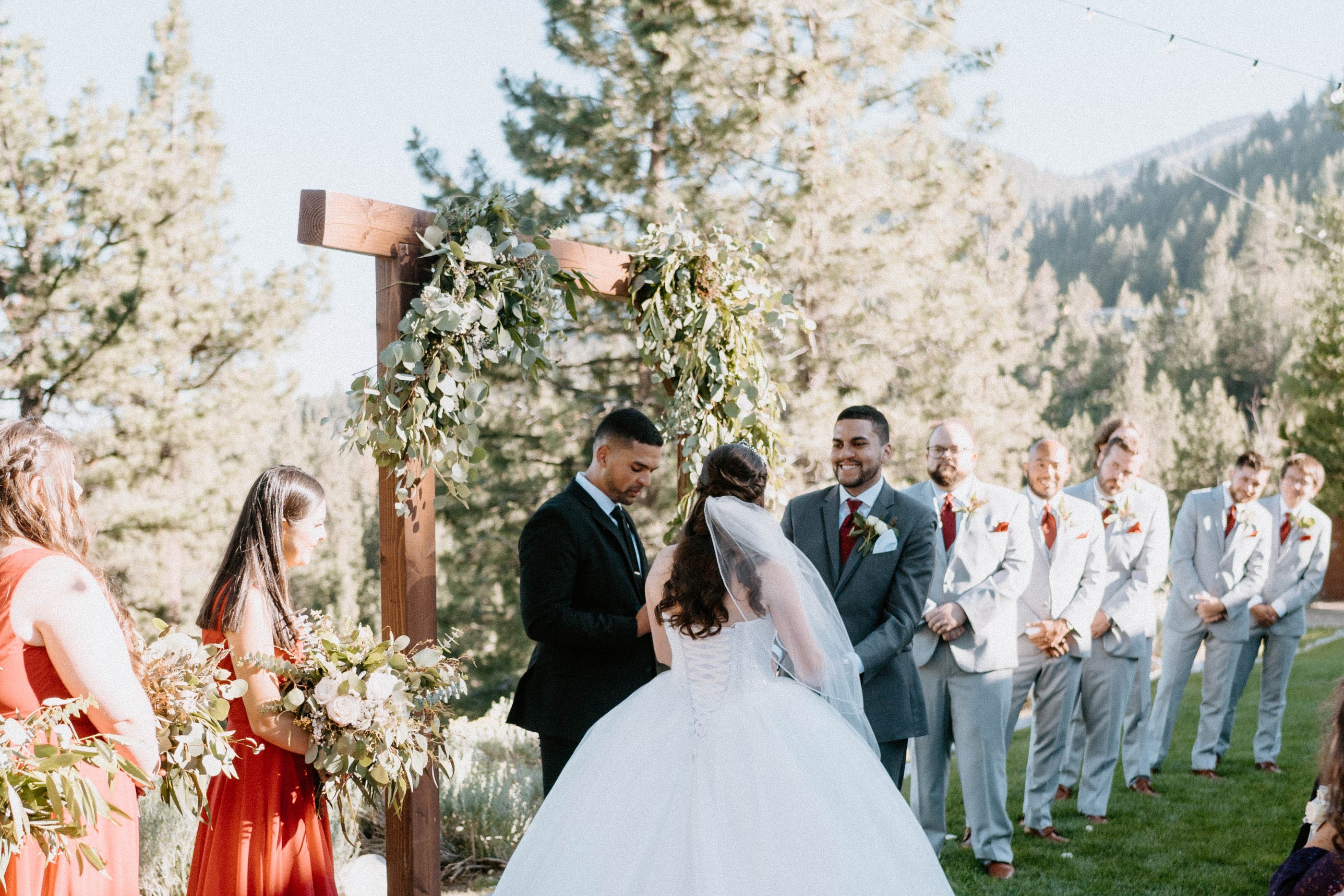 Montse and Jason at the wooden arch during their ceremony, bridesmaids in red on one side and groomsmen in grey on the other