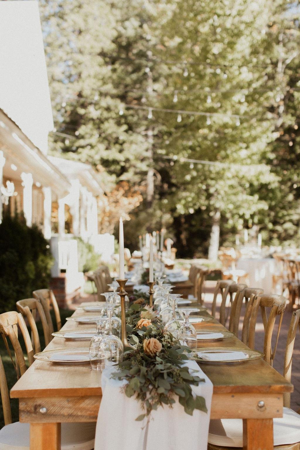 A long wooden table set with greenery and candles beside the white house, photo by Kay Kroshus Photography