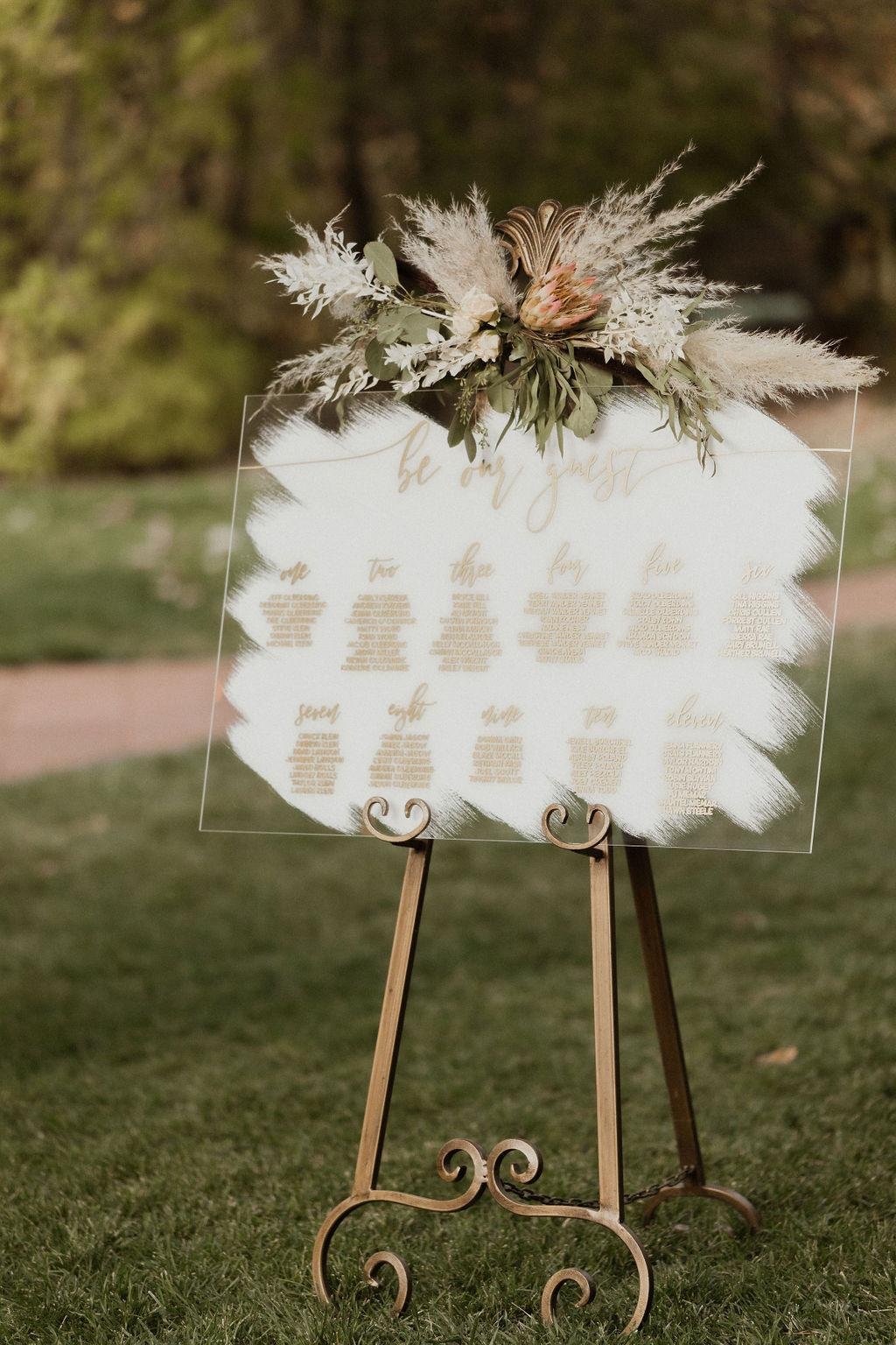 An acrylic seating chart topped with pampas grass on a brass easel, photo by Kay Kroshus Photography
