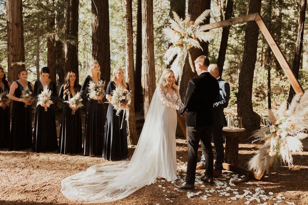Nicole and Thomas say their vows at a triangle arch of pampas grass, her bridesmaids in black beside her, photo by Kay Kroshus Photography