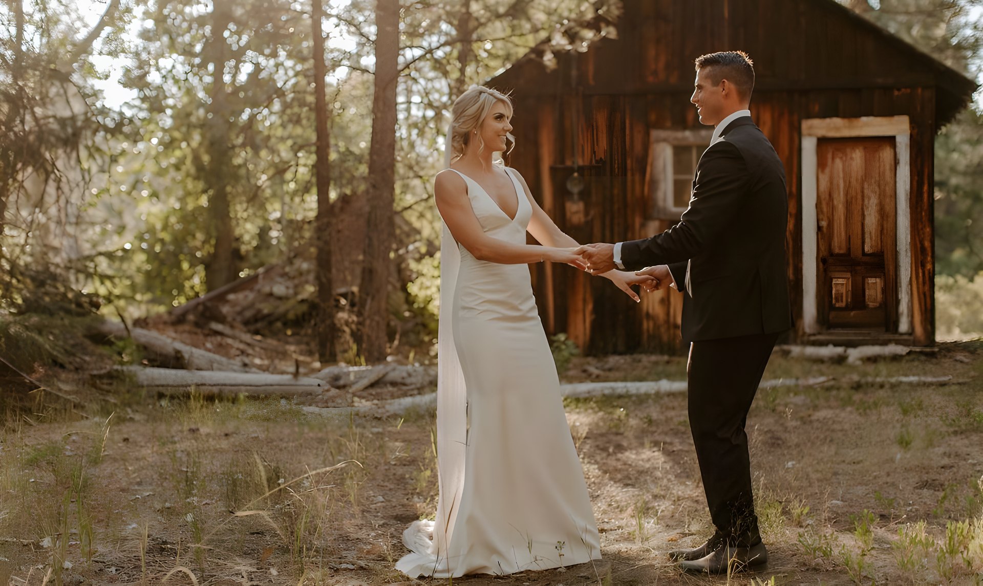 Nicole and Tim smile and hold hands in the pines in front of an old wooden cabin, photo by Taylor McCutchan Photography