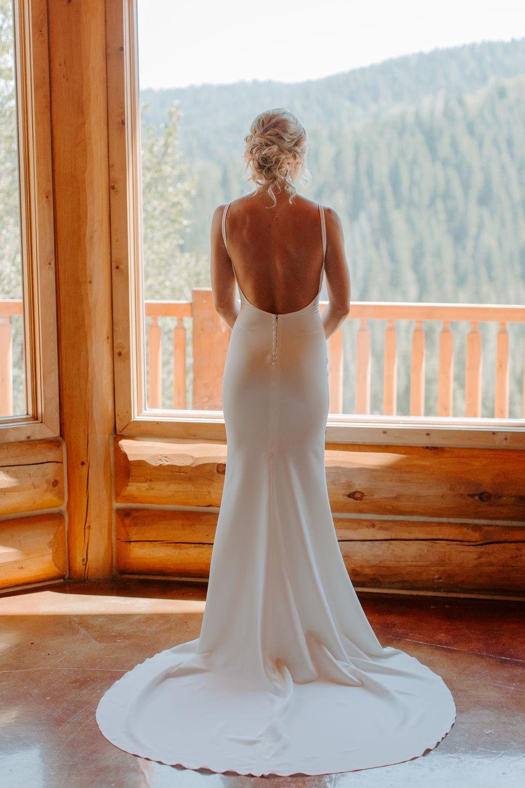 Nicole stands at the cabin window looking out at the pines in her backless gown, photo by Taylor McCutchan Photography