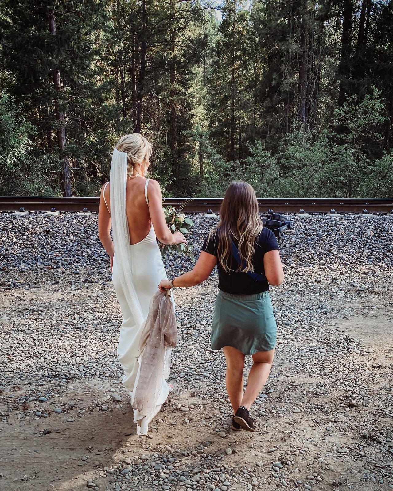 Nicole in her open back gown walks along the railroad tracks in the pines with a woman carrying her train