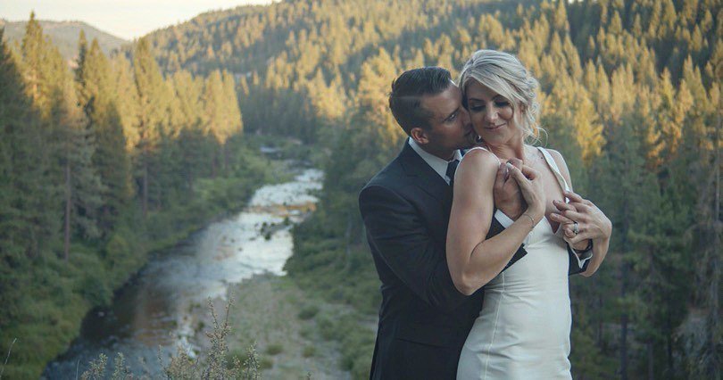 Tim holds Nicole from behind on a hillside above a river winding through the pines