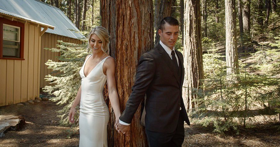 Nicole and Tim hold hands on either side of a tall pine for their first look beside a wooden cabin