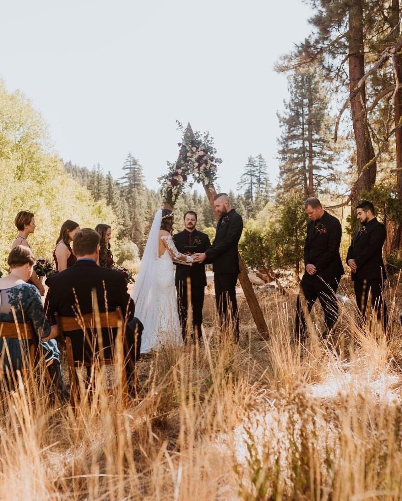 Guests watch Rachel and Chris at the arch in the tall meadow grass among the pines