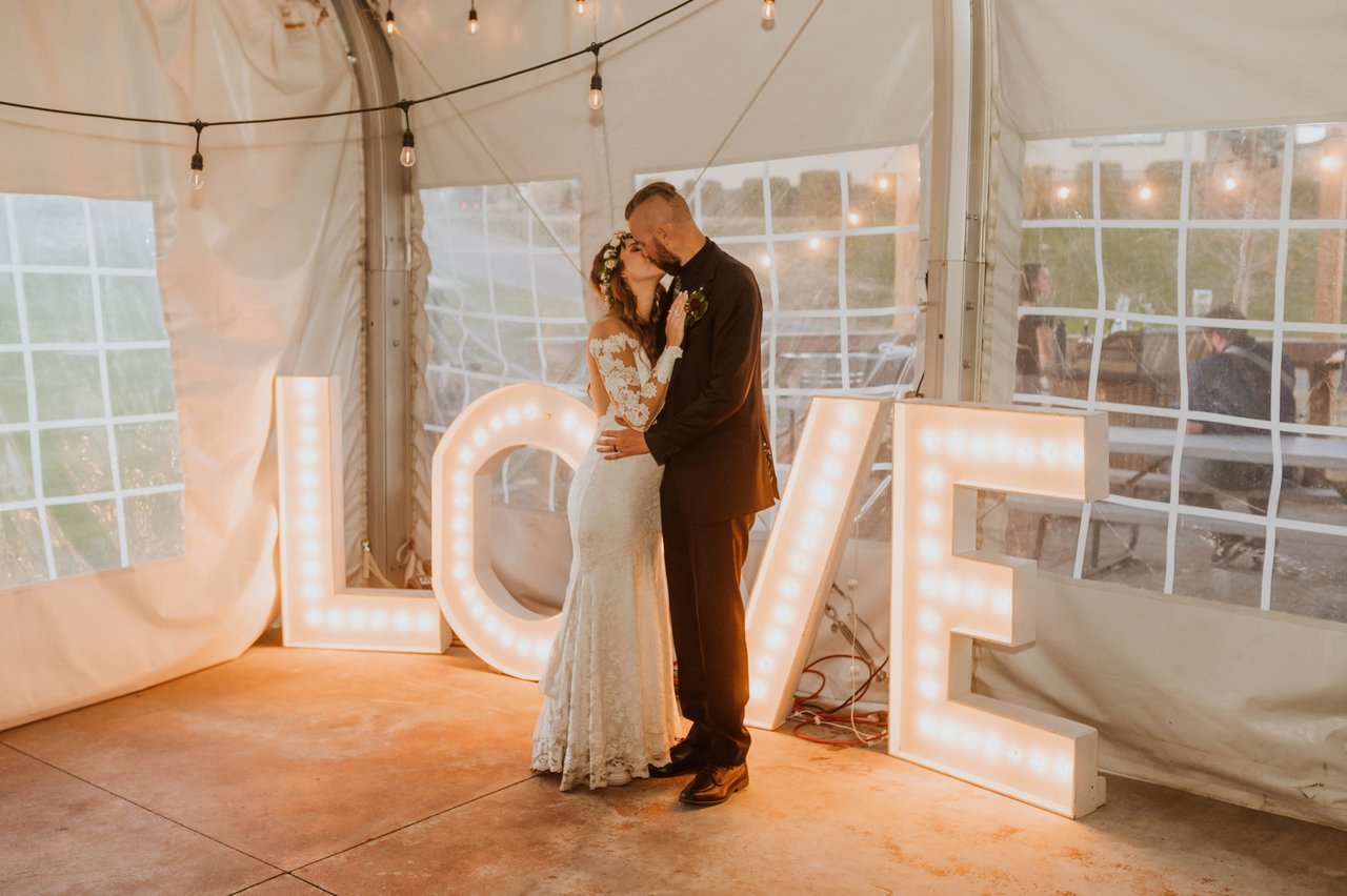 Rachel and Christopher kiss beside glowing LOVE letters in the tent
