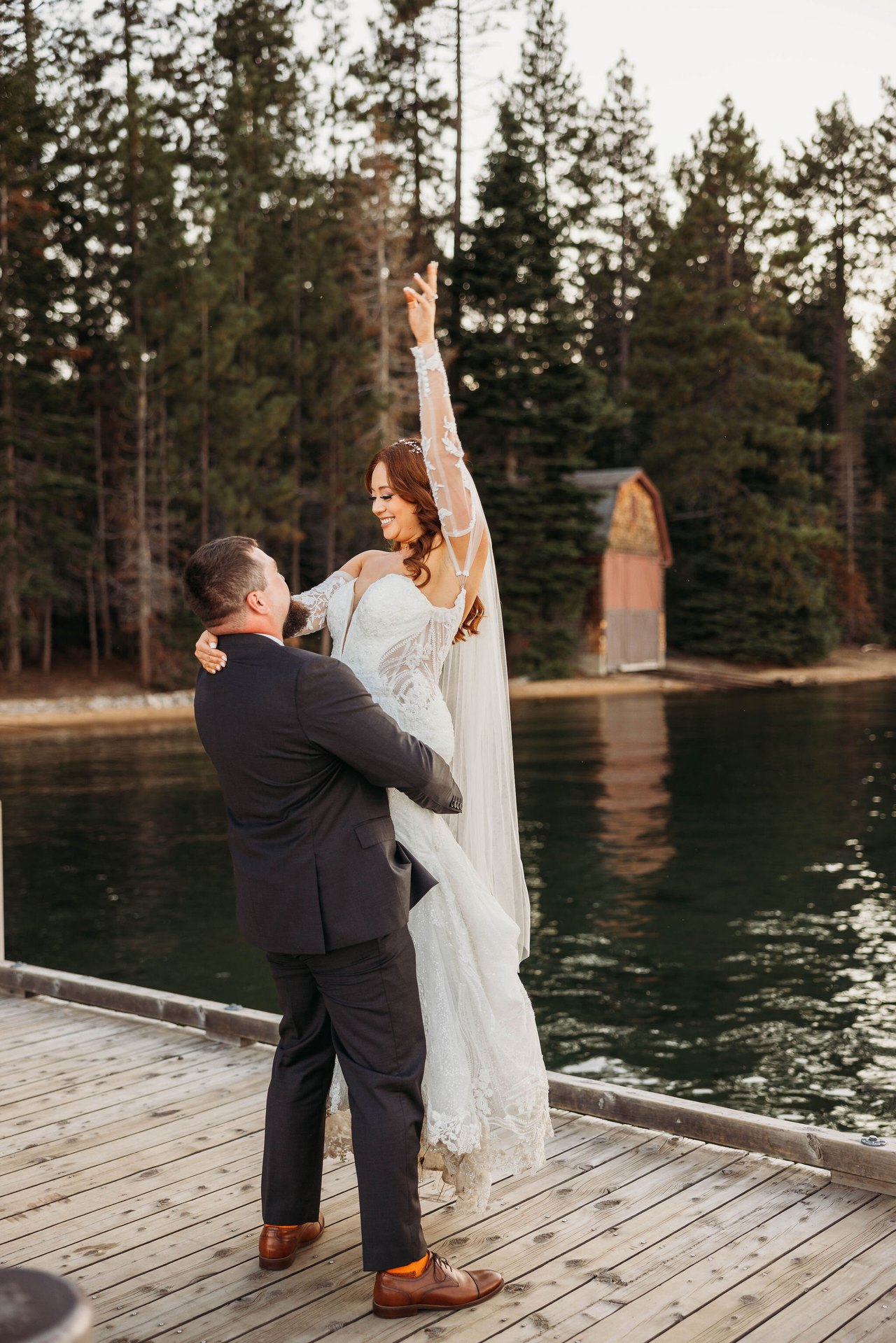 Tom lifts Remy on the dock as she raises one arm, the red boathouse behind them