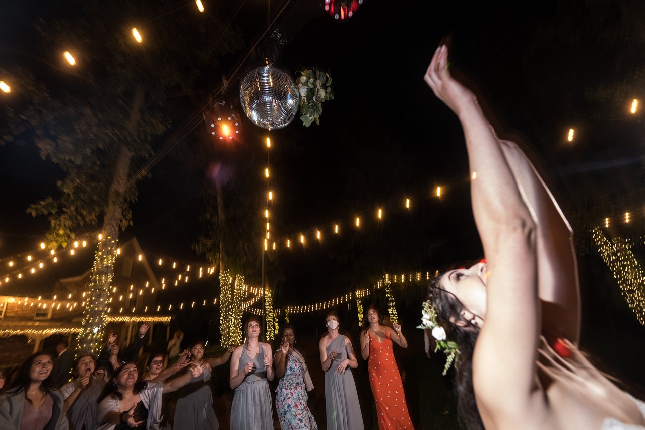 Shannon flings her bouquet over her head as her friends reach for it under the string lights