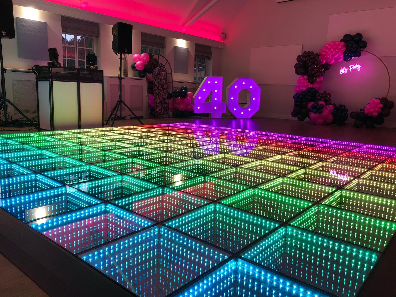A rainbow lit LED infinity dance floor in a party hall set for a 40th birthday, the DJ booth on the left